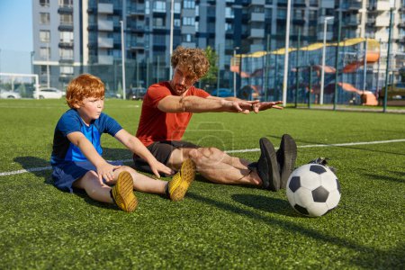 Téléchargez les photos : Père et fils concentrés faisant de l'exercice d'étirement avant de jouer au football sur l'herbe au terrain de soccer à l'extérieur - en image libre de droit