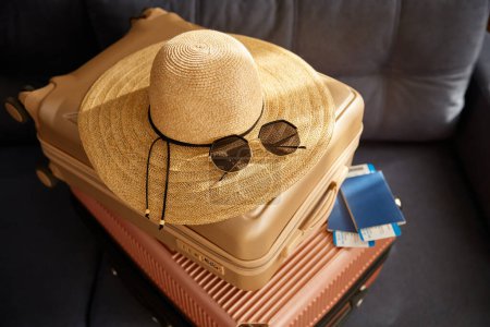 Téléchargez les photos : Valise de voyage empilée avec chapeau de plage en paille, lunettes de soleil et document de vol sur canapé en cuir. Réservation voyage, vacances d'été et concept de préparation de tour de station - en image libre de droit