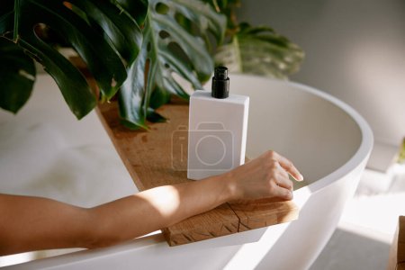 Téléchargez les photos : Bouteille de produit d'hygiène pour se baigner sur la baignoire et la main féminine. Soins du corps et des cheveux routine quotidienne, détente au spa à la maison - en image libre de droit