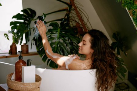 Téléchargez les photos : Superbe femme appliquant du gel moussant sur le corps pour le nettoyage tout en prenant un bain à la maison. Traitement spa et concept de beauté féminine - en image libre de droit