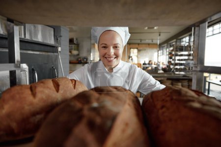 Téléchargez les photos : Portrait d'une pâtissière souriante regardant à travers une grille avec des pains fraîchement cuits. Travailleur boulangerie - en image libre de droit