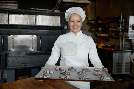 Téléchargez les photos : Joyeux boulanger souriant avec plateau de biscuits fraîchement cuits debout à la cuisine de boulangerie regardant la caméra - en image libre de droit