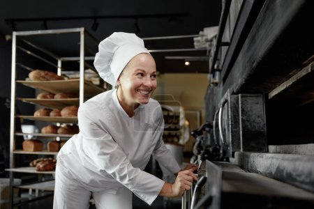 Photo for Female baker with smile on face looking at kitchen oven with bread or pastry checking and controlling baking process at food manufacturing - Royalty Free Image