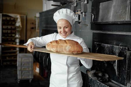 Téléchargez les photos : Une pâtissière souriante sent le pain fraîchement cuit sur une pelle en bois. Travailleur boulangerie - en image libre de droit