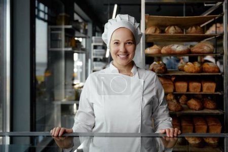 Téléchargez les photos : Portrait de belle femme souriante en uniforme debout à la table de livraison de la boulangerie avec du pain fraîchement cuit sur des supports en acier - en image libre de droit