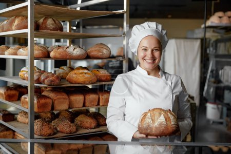 Photo for Happy woman baker chef holding bread standing near showcases with baked loaf. Bakery shop concept - Royalty Free Image