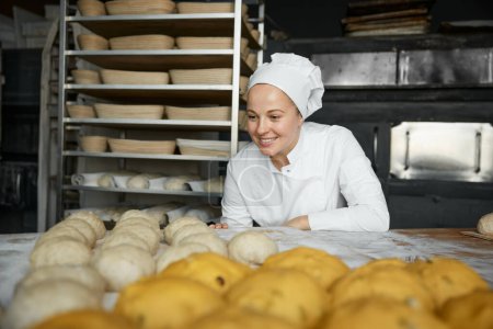 Téléchargez les photos : Satisfait femme boulangère positive à l'atelier de boulangerie artisanale. Femme chef en uniforme regardant les préparations crues de petits pains sucrés sur la table dans la farine - en image libre de droit