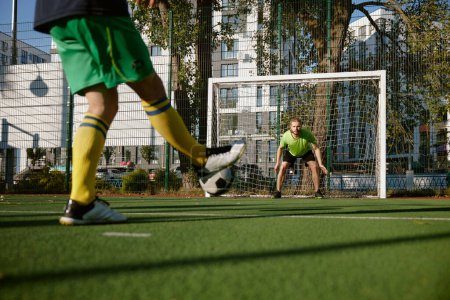 Photo for Football player aiming soccer ball trying to score goal into gate defended by goalkeeper. Active summer holiday outdoor and sport match between friends - Royalty Free Image