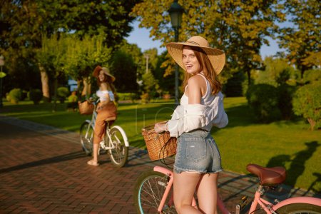 Téléchargez les photos : Deux belles filles meilleures amies marchant à vélo au parc public. Charmante jeune fleuriste féminine profitant de la compagnie de l'autre - en image libre de droit