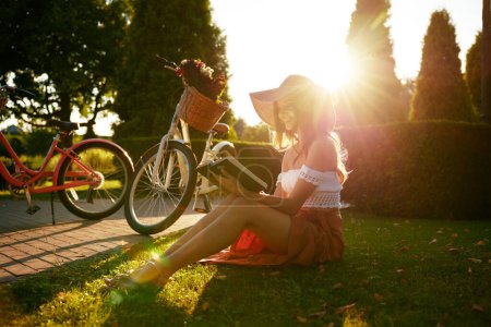 Téléchargez les photos : Charmante jeune femme lisant le livre tout en étant assis dans le parc sur la pelouse dans le rayon de lumière du soleil sur le coucher du soleil. Adolescente joyeuse relaxante profitant de la fin heureuse de la belle journée en plein air - en image libre de droit