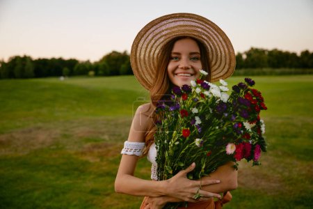 Foto de Retrato facial de una hermosa mujer hipster sosteniendo un ramo de flores silvestres de pie sobre el campo verde. Feminidad, belleza natural y concepto de inocencia - Imagen libre de derechos