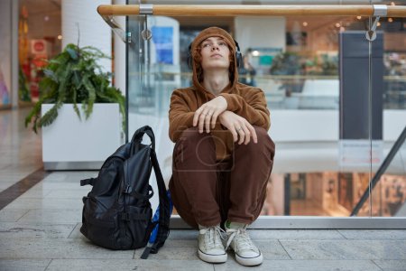 Foto de Adolescente trastornado con auriculares sentados cerca de escaleras móviles en el centro comercial. Deprimido chico serio escuchando música sintiendo emoción negativa - Imagen libre de derechos