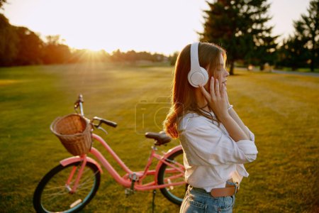 Téléchargez les photos : Jeune fleuriste hipster femme portant des écouteurs écoutant de la musique se reposer après une longue course à vélo pour les fleurs sauvages sur le terrain vert. Artisanat passe-temps et voyager à l'extérieur - en image libre de droit
