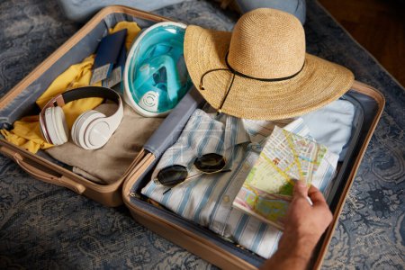 Photo for Male hands putting paper map into travel bag with summer clothing while packing stuff for travel, closeup view. Summer vacation trip adventure aspirations concept - Royalty Free Image