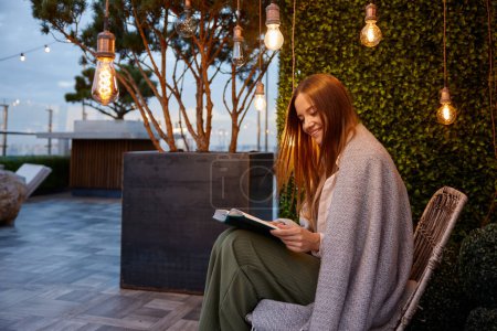Foto de Mujer joven envuelta en libro de lectura de manta caliente disfrutando de la relajación en un lugar acogedor de la terraza del hogar o la cafetería al aire libre. Sueño romántico estado de ánimo e imaginación - Imagen libre de derechos