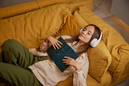 Téléchargez les photos : Une jeune femme s'est endormie en lisant un livre et en écoutant de la musique dans un casque. Vue aérienne sur une belle fille charmante dormant sur un canapé à la maison - en image libre de droit