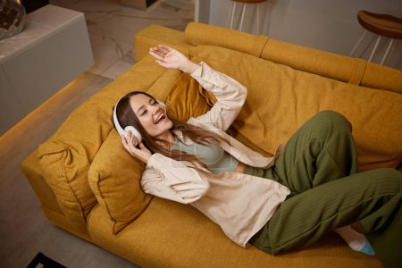 Foto de Feliz mujer joven relajada escuchando música en los auriculares en casa. Milenial mujer acostada en cómodo sofá - Imagen libre de derechos