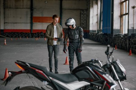 Photo for Student motorcyclist and instructor discussing rout of future exam race. Two men standing together looking at checklist at indoor motordrome of driver training center - Royalty Free Image