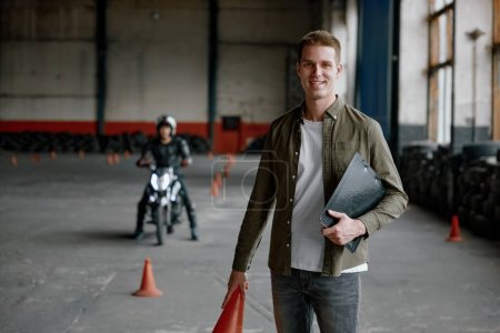 Photo for Portrait of happy smiling instructor of driving school at training motordrome with motorcyclist student riding motorbike on background - Royalty Free Image