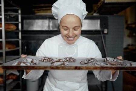 Photo for Happy smiling baker with tray of freshly baked cookies standing at bakery kitchen looking at camera - Royalty Free Image