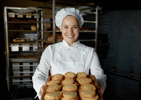 Foto de Mujer chef con bandeja de apetito deliciosa pastelería de pie en la cocina de la panadería. Preparación de galletas de avena - Imagen libre de derechos