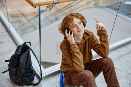 Téléchargez les photos : Adolescent garçon se détendre au centre commercial assis sur le sol écouter de la musique et profiter de temps libre seul. Concept d'émotion insouciante - en image libre de droit