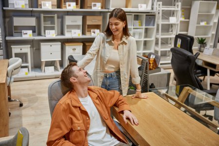 Photo for Young family couple choosing computer desk at modern furniture showroom. Attractive wife making surprise present for husband buying table for home office workplace - Royalty Free Image