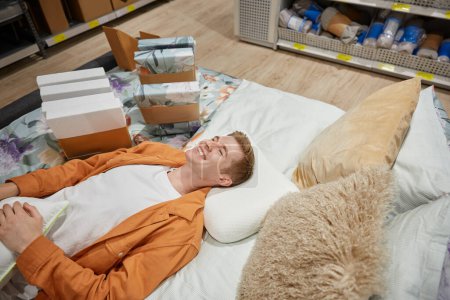 Photo for Handsome relaxed man lying on bed with pillow at shop store department. Young guy enjoying shopping choosing new bedding for his home bedroom - Royalty Free Image