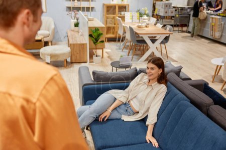Photo for Young married couple looking for sale prices on new sofa couch at furniture store. Attractive wife finding best divan offering husband to buy - Royalty Free Image