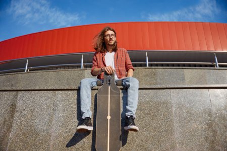 Téléchargez les photos : Portrait de skateboarder masculin hipster souriant et élégant bénéficiant d'une bonne formation au parc urbain de la ville. bel homme assis sur parapet avec longboard - en image libre de droit