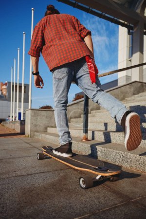 Téléchargez les photos : Hipster homme à cheval skateboard sur la rue de la ville. Casual gars avec un pied placé sur longboard et pousser un autre de la route asphaltée. Concept de style de vie sportif - en image libre de droit
