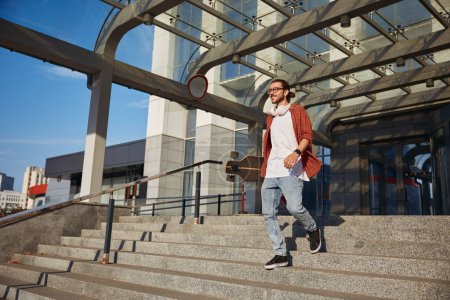 Photo for Young hipster skateboarder walking down steps with longboard in hand. Urban hobby and weekend free time activity concept - Royalty Free Image