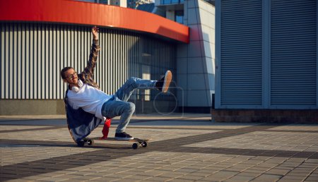 Photo for Excited skilled hipster guy making stunt on skateboard riding fast on urban street. Fashionably dressed teenager boy skater doing handstand trick. Sport hobby of young people concept - Royalty Free Image