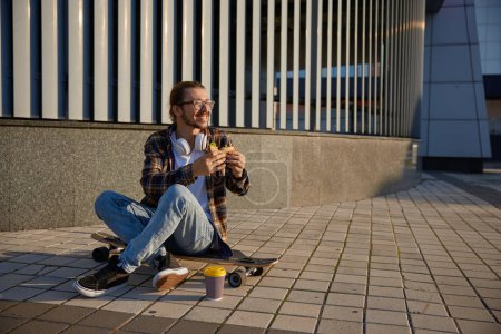 Foto de Hipster hombre con ropa de moda de moda con longboard comer sándwich. Cultura urbana y deporte urbano - Imagen libre de derechos