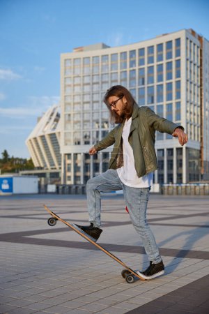 Photo for Skilled hipster guy making stunt on skateboard riding fast on urban street. Fashionably dressed teenager boy skater doing handstand trick. Sport hobby of young people concept - Royalty Free Image