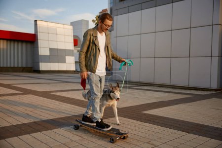 Photo for Hipster guy in casual clothing skateboarding and walking dog moving together through city square. Pet owner and loving puppy enjoying physical activity during calm evening at downtown district - Royalty Free Image