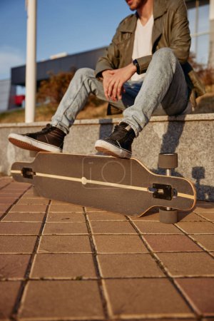 Photo for Cropped shot of relaxed hipster skateboarder with focus on foot placed on board while sitting on sidewalk parapet. Sports recreation at public park. Street style and youth culture - Royalty Free Image