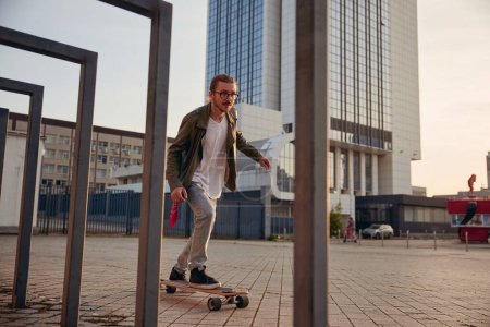 Téléchargez les photos : Cool type banlieue sur skateboard sur fond de ville moderne. Jeune bel homme conduisant longboard garder l'équilibre pendant la course de vitesse - en image libre de droit