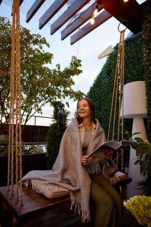 Foto de Mujer joven envuelta en una manta cálida disfrutando de la relajación en un lugar acogedor de la terraza del hogar o la cafetería al aire libre. Sueño romántico estado de ánimo e imaginación - Imagen libre de derechos