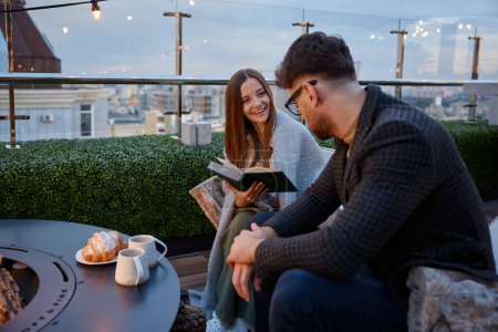 Photo for Young girlfriend reading for boyfriend while couple resting at roof top cafe. Excited female sharing impression of favorite book. Young family leisure activity in evening - Royalty Free Image