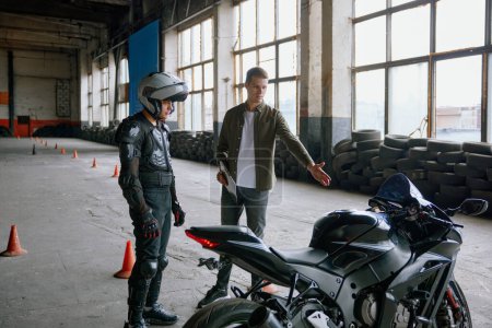 First lesson at motorcycle school. Instructor describing technical characteristics of motorcycle and safety traffic rules to student wearing protective suit and helmet