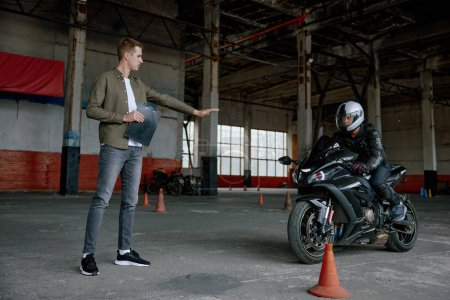 Téléchargez les photos : Jeune homme motocycliste apprendre à faire du vélo à l'école privée manœuvrant entre les cônes à moto intérieure - en image libre de droit