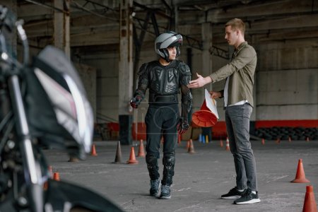 Photo for Student motorcyclist and instructor discussing rout of future exam race. Two men standing together looking at checklist at indoor motordrome of driver training center - Royalty Free Image