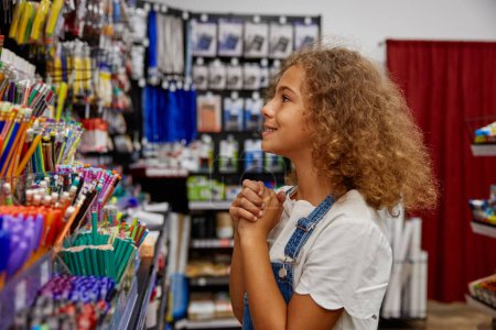 Téléchargez les photos : Petite écolière choisissant stylo au magasin de papeterie tout en se préparant pour la nouvelle année scolaire - en image libre de droit