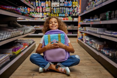 Photo for Portrait of happy girl hugging new colorful schoolbag while sitting on floor of stationery store. Back to school, preparation for study and knowledge receiving process - Royalty Free Image