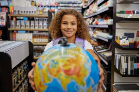 Photo for Little girl playing with school earth globe while shopping education stationery. Positive female student preparing for studying. Purchases with sale discount - Royalty Free Image