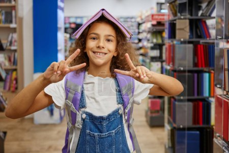 Photo for Portrait of happy girl child with opened diary on head gesturing peace victory sign looking at camera holding schoolbag on back while standing between stationery store. Shopping time concept - Royalty Free Image