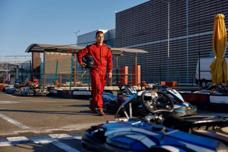 Well-equipped go-kart driver wearing overalls and holding helmet walking to car before race at starting line on motor racing track. Automotive sports activity, carting championship