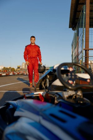 Photo for Confident go-kart driver wearing overalls and holding helmet walking to car before race at starting line on motor racing track. Automotive sports activity, carting championship - Royalty Free Image