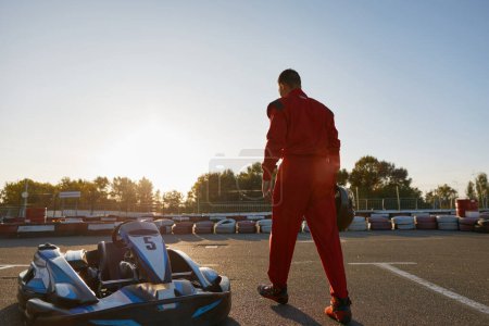 Photo for Confident go-kart driver wearing overalls and holding helmet walking to car before race at starting line on motor racing track. Automotive sports activity, carting championship - Royalty Free Image